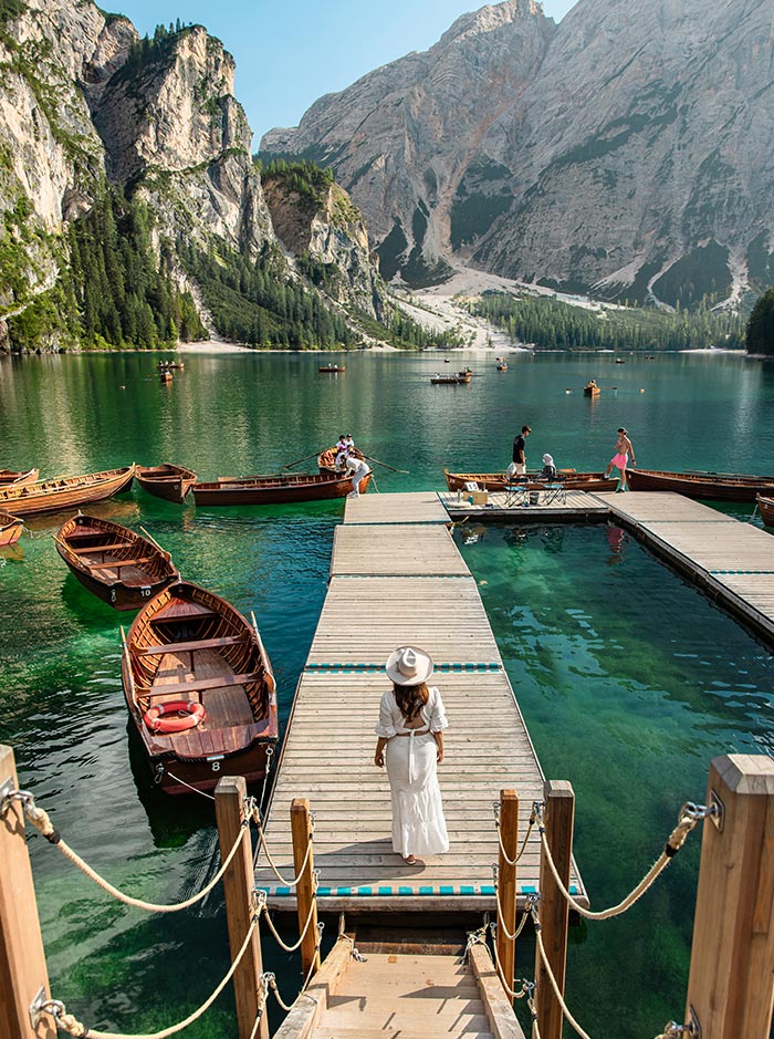 This image captures a serene scene at the dock of Lago di Braies (Pragser Wildsee) in the Dolomites. A woman in a white dress and hat stands on the wooden pier, surrounded by traditional wooden boats on the clear, emerald-green lake. In the background, steep, forested cliffs and towering mountains rise dramatically above the water. Other visitors can be seen preparing for boat rides, adding to the tranquil, yet lively atmosphere of this beautiful alpine destination.