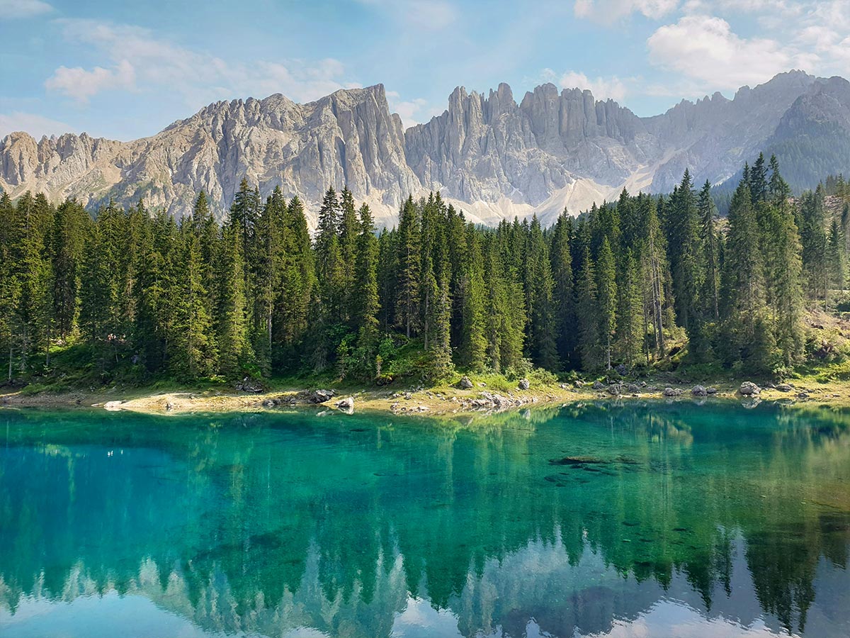 This image features the crystal-clear turquoise waters of Lago di Carezza (Karersee) in the Dolomites, perfectly reflecting the surrounding dense forest and towering mountain peaks in the background. The peaceful lake is framed by tall trees and rugged cliffs, creating a stunning contrast between the vibrant blue water and the natural alpine scenery. The majestic mountain range adds to the serene, postcard-like beauty of this iconic Dolomite location.