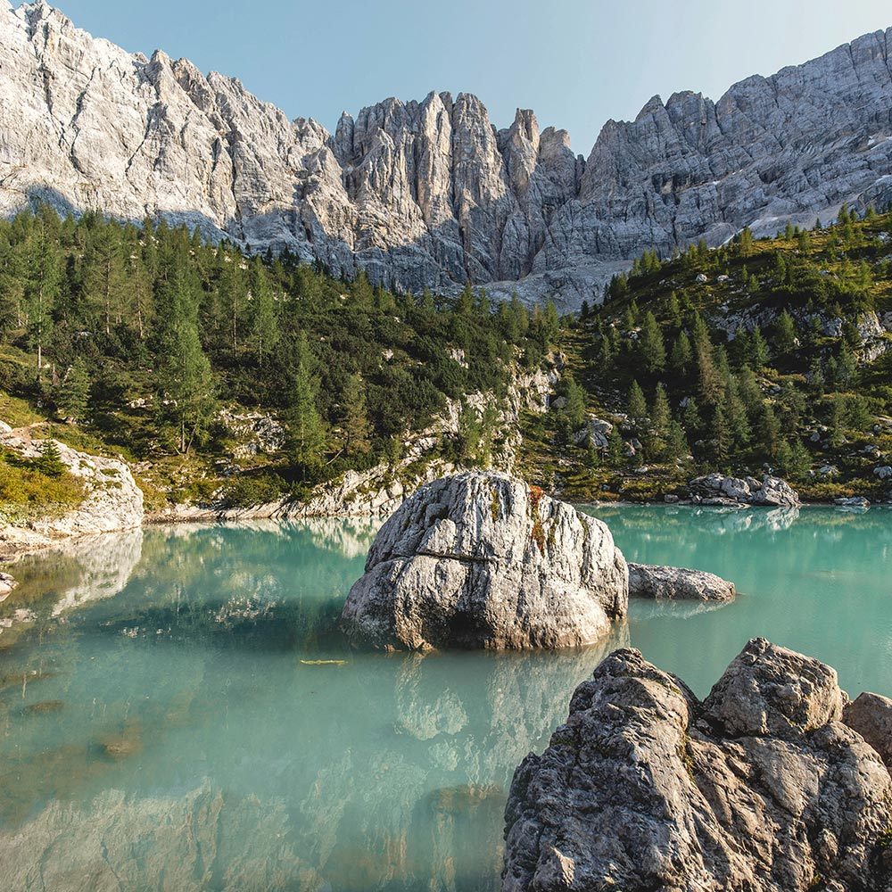 This image showcases the striking turquoise waters of Lago di Sorapis in the Dolomites, surrounded by rugged, towering cliffs and lush greenery. Large boulders sit in the calm, reflective water, while alpine trees dot the landscape around the lake. The dramatic rock formations and the vivid color of the lake create a breathtaking contrast, making this alpine scene both serene and awe-inspiring.