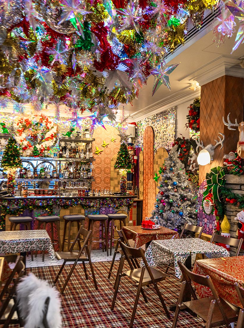 This image shows the festive interior of the Miracle Christmas Bar at the Henrietta Hotel, decorated with vibrant holiday colors. The ceiling is adorned with shimmering tinsel, stars, and ornaments, creating a dazzling overhead display. The bar area features mini Christmas trees and garlands, while the tables are covered with colorful, holiday-patterned cloths. A silver Christmas tree and various holiday decorations fill the cozy space, enhancing the warm, celebratory atmosphere.