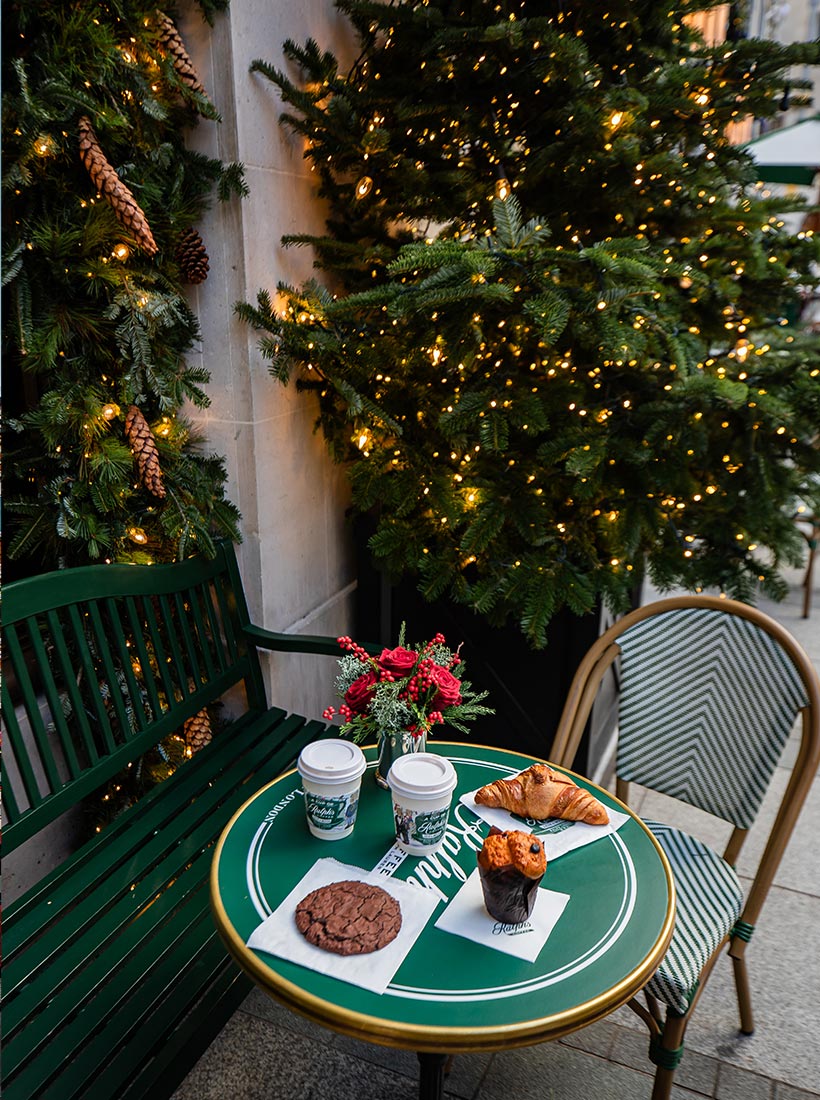 This image captures a festive outdoor scene at Ralph's Coffee on New Bond Street in London. A small green table is set with pastries, including a croissant, muffin, and cookie, alongside two cups of coffee. The table is adorned with a small bouquet of red roses and greenery, complementing the holiday decor. Surrounding the seating area are twinkling Christmas lights on lush evergreen garlands and trees, creating a cozy and inviting atmosphere for enjoying a warm treat during the holiday season.