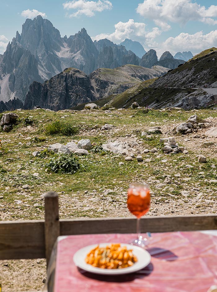 This image shows a plate of pasta and a glass of Aperol spritz on a table with an awe-inspiring view of jagged mountain peaks in the Dolomites. The rocky, rugged terrain and distant cliffs create a dramatic backdrop, while the outdoor dining setup suggests a peaceful meal at a mountain refuge. The combination of delicious food and breathtaking scenery creates a perfect alpine dining experience.