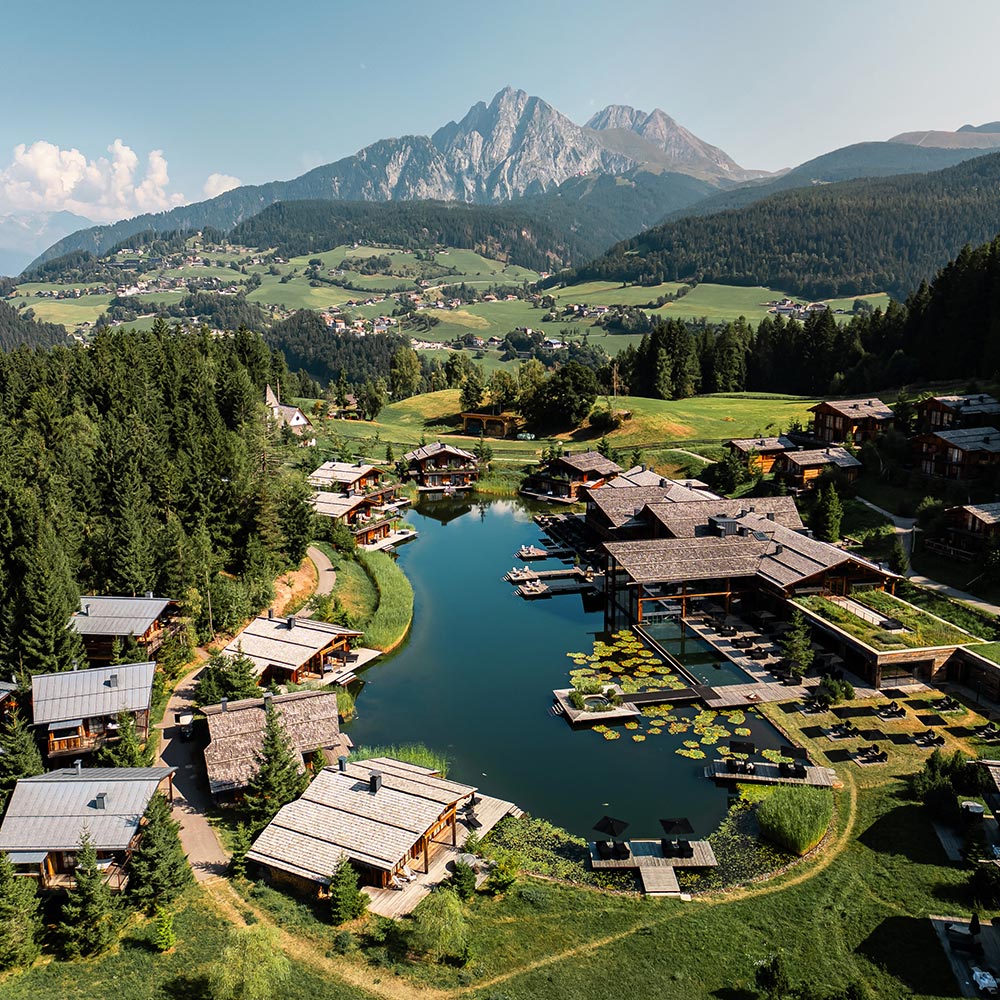 This image features an aerial view of a luxury lodge retreat centered around a peaceful lake, surrounded by dense forests and green rolling hills, with majestic mountains in the background. The wooden lodges are arranged around the lake, with docks and seating areas extending over the water, offering a blend of rustic charm and modern comfort. The expansive landscape showcases the beauty of nature combined with the serenity of this high-end mountain getaway.






