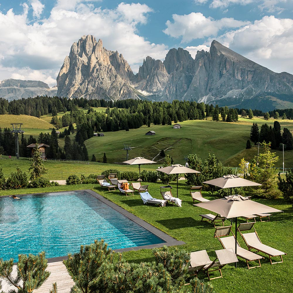 This image showcases a serene outdoor pool surrounded by lounge chairs and umbrellas, set against a backdrop of rolling green meadows and towering rocky peaks of the Dolomites. The scene exudes tranquility and luxury, blending natural beauty with comfortable leisure spaces. The rugged mountain range in the distance contrasts beautifully with the lush greenery and the inviting poolside setting.






