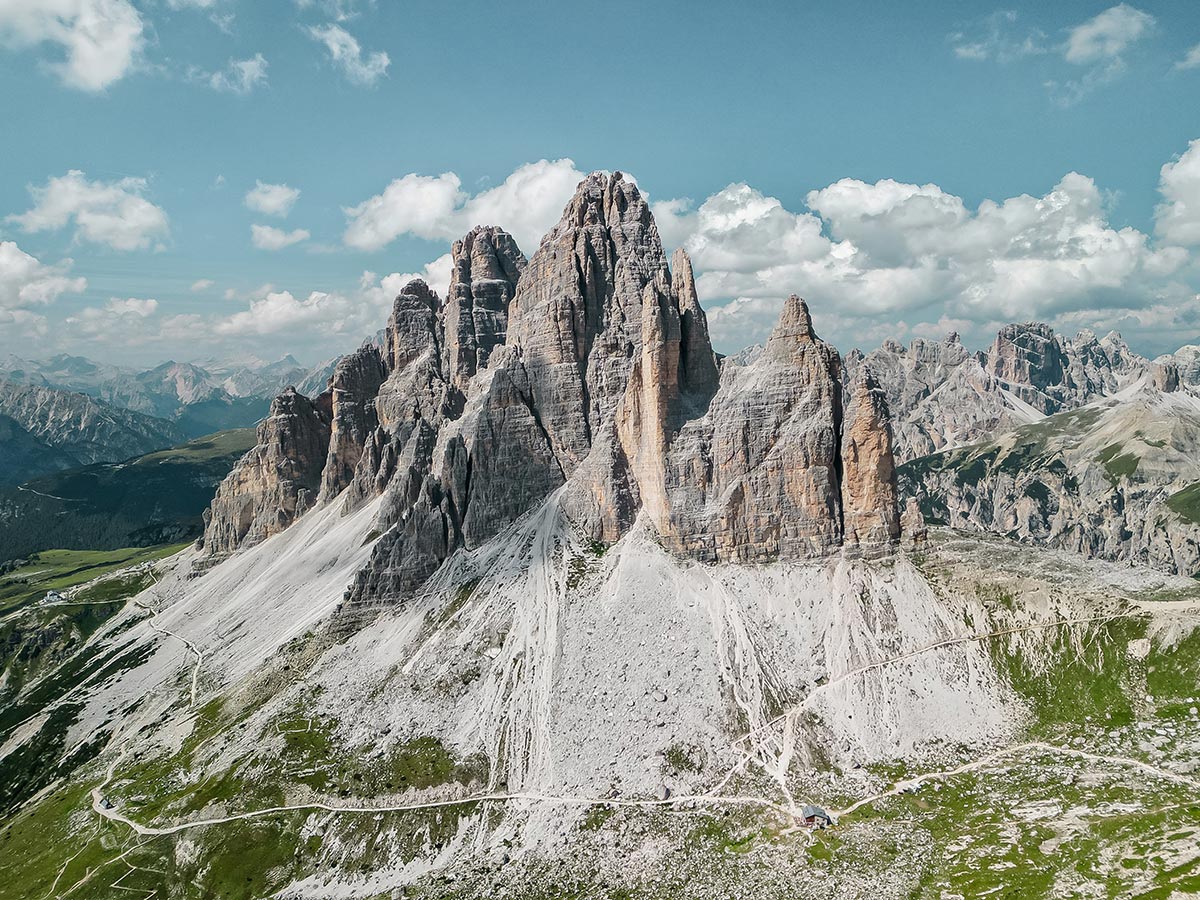 This image showcases the majestic Tre Cime di Lavaredo, three towering rock formations in the Dolomites. The jagged, rocky peaks rise dramatically above a rugged landscape of steep slopes, gravel paths, and grassy patches. The expansive view reveals a network of hiking trails that wind through the area, highlighting the impressive scale of the mountains against a backdrop of distant peaks and a partly cloudy sky. This iconic Dolomite landmark is a popular destination for adventurers and nature enthusiasts.