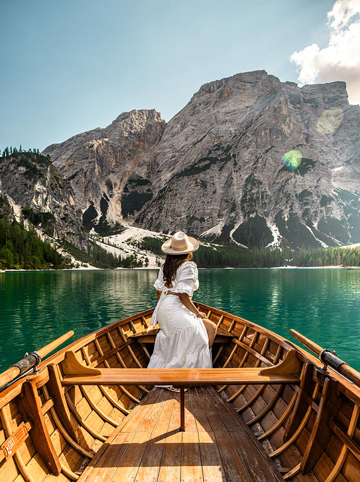 This image features a woman in a white dress and hat seated at the bow of a wooden boat, gazing at the towering mountains that rise above the turquoise waters of Lago di Braies (Pragser Wildsee) in the Dolomites. The peaceful lake reflects the surrounding rocky cliffs and dense forests, creating a serene and majestic atmosphere. The image captures a moment of tranquil exploration and connection with nature in this stunning alpine setting.