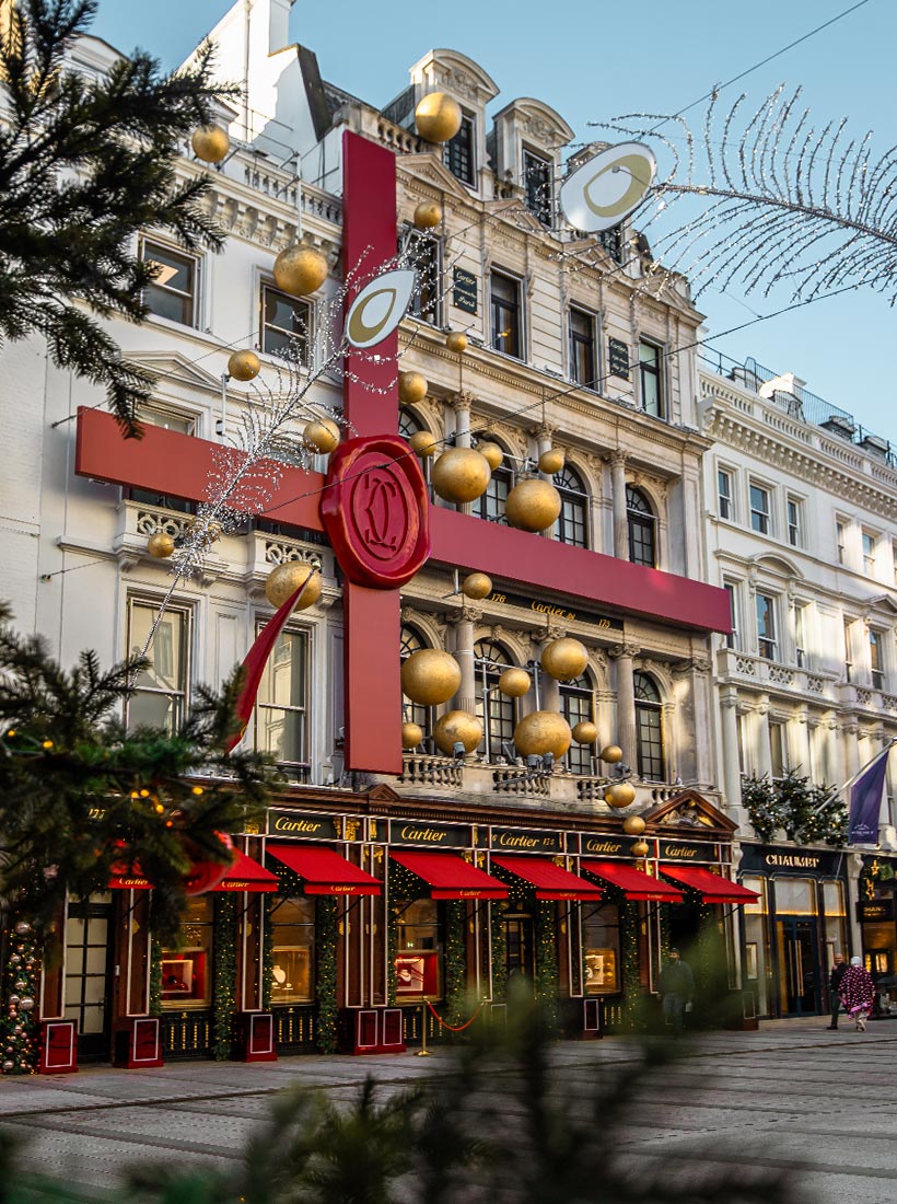 This image shows the Cartier store on Old Bond Street in London, decorated for Christmas with a massive red ribbon wrapped around the building, resembling a gift. The iconic Cartier logo is displayed as a large wax seal at the center of the ribbon. Gold ornaments and festive decorations are scattered across the facade, while the storefront is adorned with red awnings and holiday lights, creating a luxurious holiday atmosphere. The scene is framed by greenery, adding to the festive charm of the street.