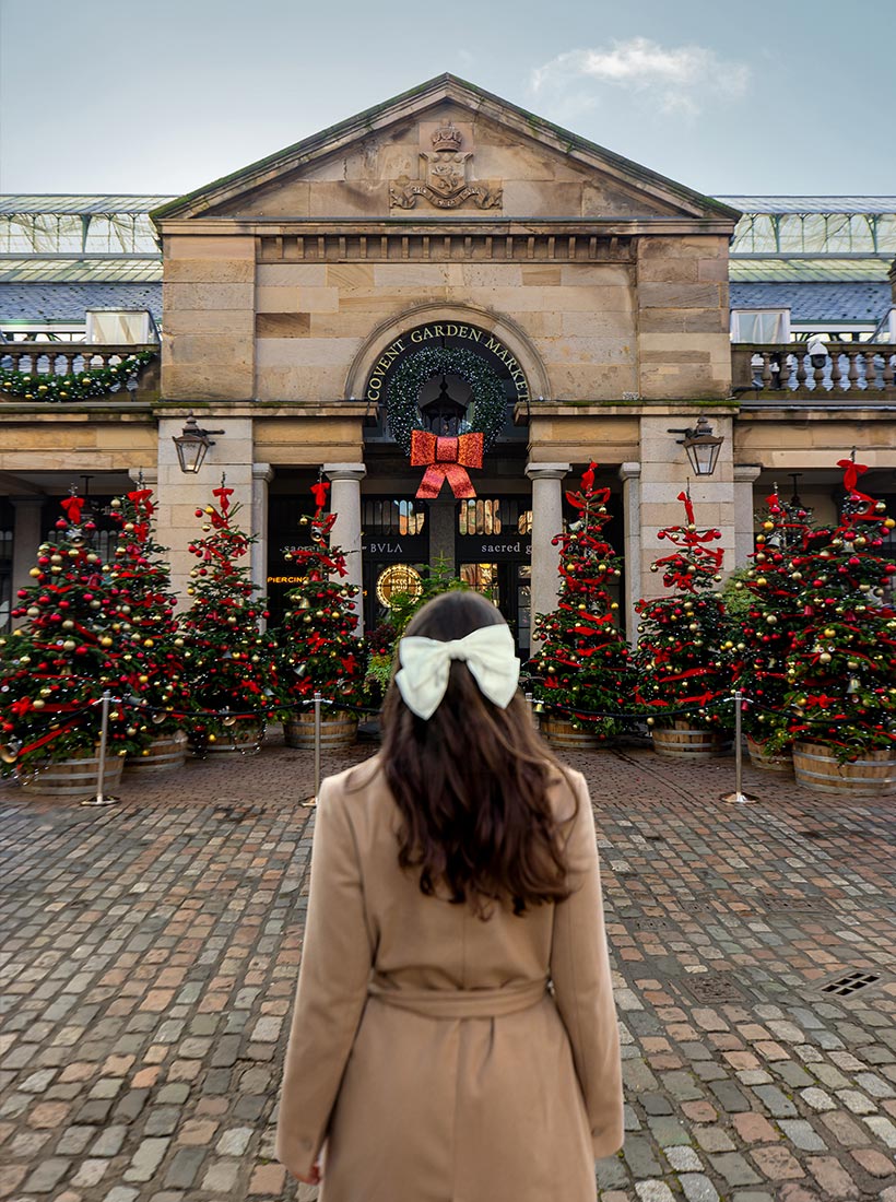 This image shows a person with long hair and a large white bow standing in front of the Covent Garden Market in London, which is decorated for Christmas. The entrance is adorned with a large wreath and a red bow, and several Christmas trees with red and gold ornaments line the path. The cobblestone street and festive atmosphere highlight the holiday spirit.