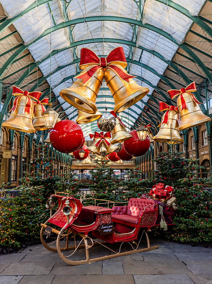 This image features a festive holiday display in Covent Garden, London, showcasing a decorated sleigh surrounded by Christmas trees adorned with lights and presents. Above the scene, large golden bells with red bows, oversized red ornaments, and glittering decorations hang from the ceiling of the market hall. The sleigh, with a sign that reads "Santa's gone shopping," adds to the cheerful, whimsical atmosphere of this Christmas setup.