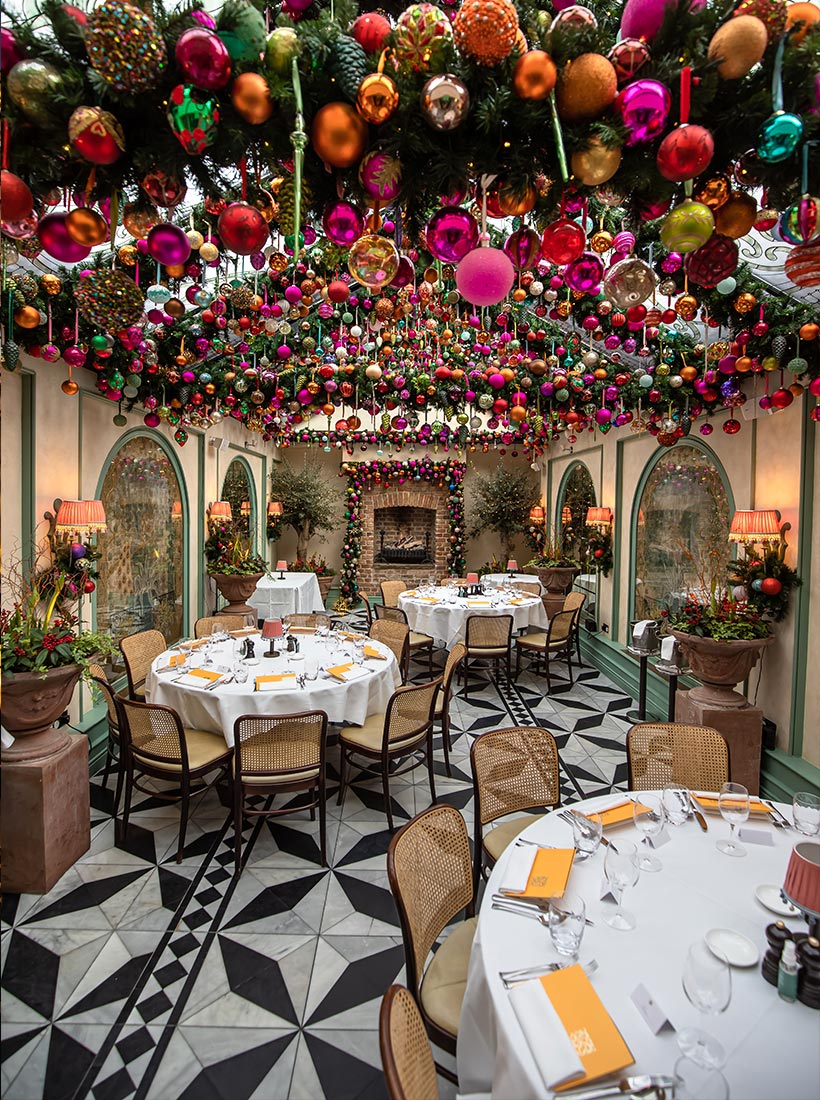 This image captures a beautifully festive dining room at Daphne's Restaurant in London, decorated for the Christmas season. The ceiling is adorned with a vibrant arrangement of hanging ornaments in shades of pink, gold, green, and red, creating a striking overhead display. The round tables are elegantly set with white tablecloths, glassware, and folded napkins, surrounded by wooden chairs with woven backs. A cozy fireplace, flanked by decorative greenery and festive accents, adds to the warm and inviting atmosphere of this holiday setting.