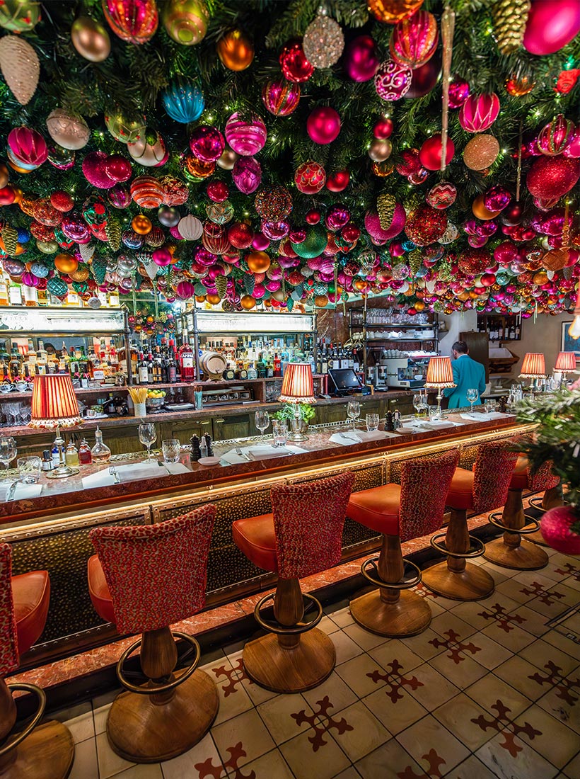 This image features the festive bar area of Daphne's Restaurant in London, lavishly decorated for Christmas. The ceiling is densely covered with a vibrant array of hanging ornaments in pink, gold, green, and red, creating a dazzling display of holiday cheer. The bar itself is elegant, with red patterned chairs, table settings, and soft glowing lamps adding warmth. The space is set for service, with neatly arranged glassware and cutlery, inviting guests to enjoy the festive atmosphere.