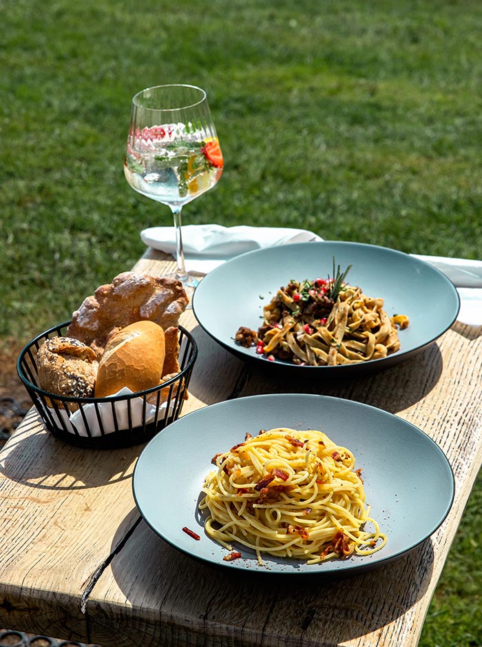 This image shows a wooden table outdoors, featuring two plates of pasta: one with spaghetti topped with crispy bacon and the other with a rich, mixed pasta dish garnished with herbs. A basket of assorted bread rolls and a glass of a refreshing drink, garnished with a slice of strawberry, sit alongside the plates. The lush green grass in the background suggests a peaceful outdoor dining experience, possibly at a mountain retreat or lodge.