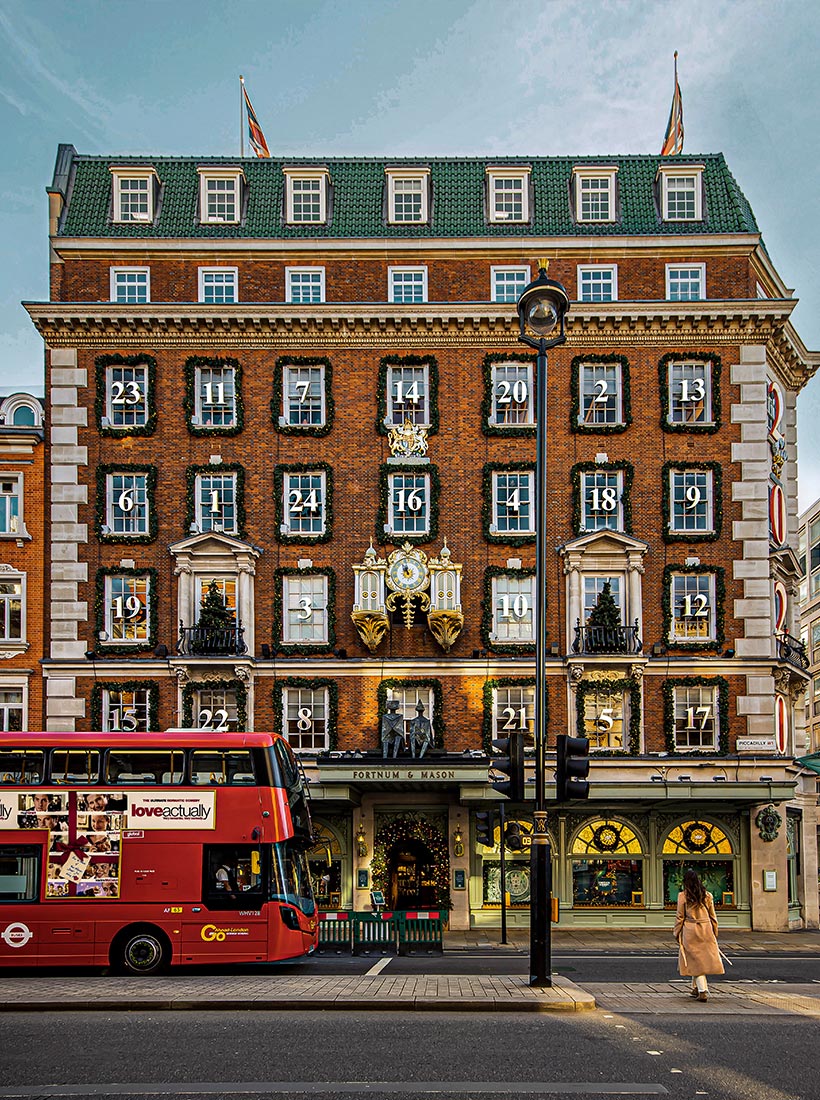 This image captures the festive facade of the Fortnum & Mason building in London, decorated as a giant advent calendar for the Christmas season. Each window is framed with garlands and numbered, with numbers 1 through 24 spread across the building. A red double-decker bus with an advertisement for "Love Actually" passes in front, and a person in a beige coat walks across the street, adding a dynamic element to the scene. The iconic Fortnum & Mason clock is visible above the entrance.