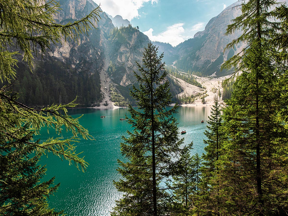 This image captures a scenic view of Lago di Braies (Pragser Wildsee) framed by lush, green trees. The turquoise water of the lake contrasts beautifully with the surrounding steep cliffs and rocky mountains. A few small boats are visible on the calm water, enhancing the peaceful atmosphere. The perspective from the forest trail offers a secluded glimpse of this stunning alpine lake, showcasing the natural beauty of the Dolomites.