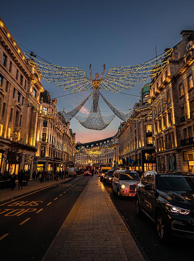 This image shows a festive holiday display on Regent Street in London during the Christmas season. Illuminated angel-shaped decorations with golden wings span across the street, creating a warm glow against the dusk sky. The street is lined with grand buildings, also adorned with lights, as cars and pedestrians move through the scene below.