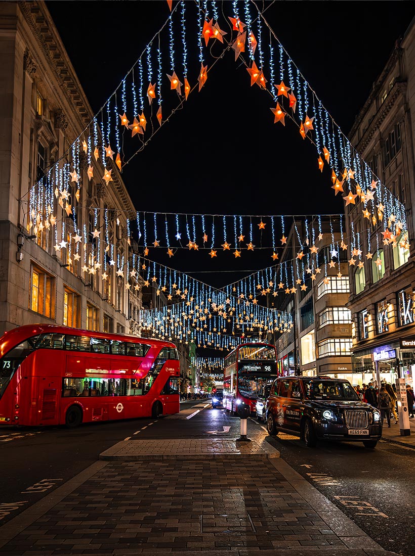 This image depicts Oxford Street in London at night, beautifully illuminated with Christmas lights. Hanging above the bustling street are strings of glowing stars and icicle-like lights, casting a festive glow over the scene. Red double-decker buses and black taxis move along the street, with shoppers visible along the brightly lit storefronts. The vibrant decorations create a magical holiday atmosphere in one of London’s most famous shopping districts.