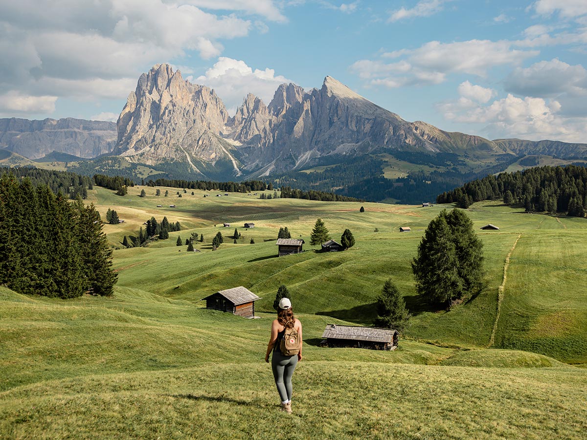 This image showcases a hiker walking through the rolling green meadows of Alpe di Siusi (Seiser Alm) in the Dolomites, with scattered wooden huts dotting the landscape. In the distance, the towering, rugged peaks of the Dolomites dominate the horizon under a partly cloudy sky. The expansive and peaceful scene emphasizes the natural beauty of the alpine region, blending wide open spaces with dramatic mountain views, perfect for outdoor exploration and adventure.