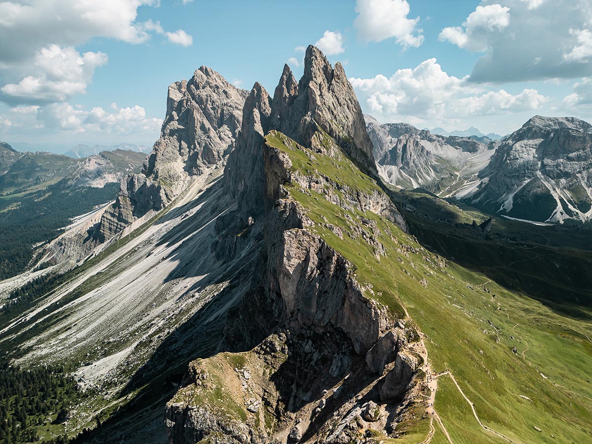 This image captures a dramatic mountain landscape featuring the sharp, jagged peaks of Seceda in the Dolomites. The towering rock formations are highlighted by steep, grassy slopes that give way to rocky cliffs, with winding trails visible along the ridges. The vast, rugged terrain stretches into the distance, creating a breathtaking scene of natural beauty, framed by a partly cloudy sky.