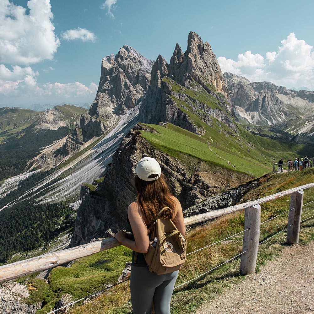 This image captures a hiker gazing at the dramatic jagged peaks of Seceda in the Dolomites from a scenic viewpoint. The sharp rock formations rise above the steep, grassy slopes and rocky terrain, creating a striking contrast. A wooden fence runs along the viewpoint, where other hikers can also be seen admiring the breathtaking landscape. The clear sky and sunlight illuminate the expansive mountain range, showcasing the natural beauty and ruggedness of this iconic Dolomite location.