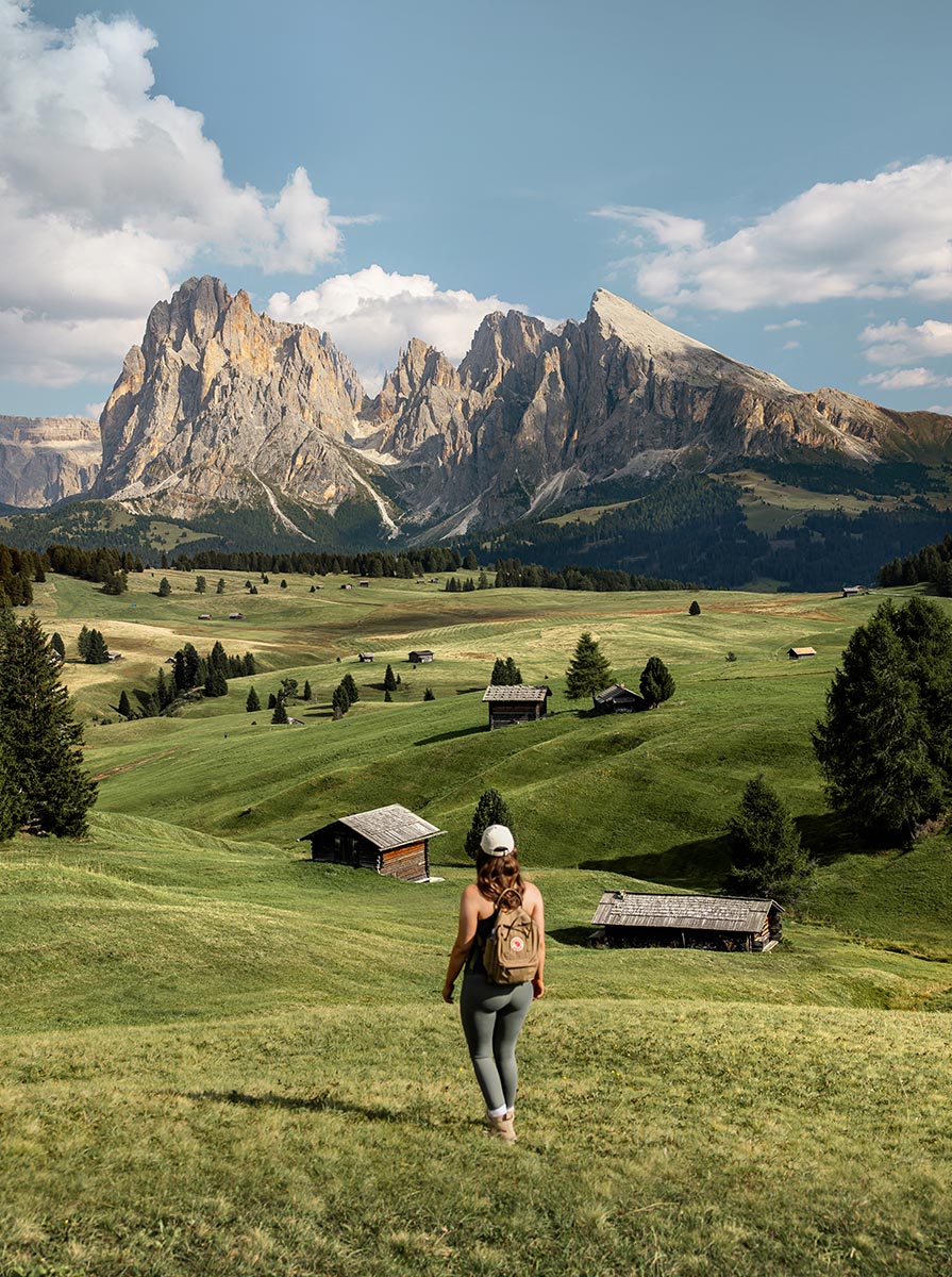 This image shows a hiker walking through the vast green meadows of Alpe di Siusi in the Dolomites, surrounded by scattered wooden huts and trees. The imposing mountain peaks rise majestically in the background under a partly cloudy sky. The hiker's backpack and outdoor gear suggest an adventurous journey through this serene, picturesque landscape, capturing the beauty and tranquility of nature in this iconic alpine region.