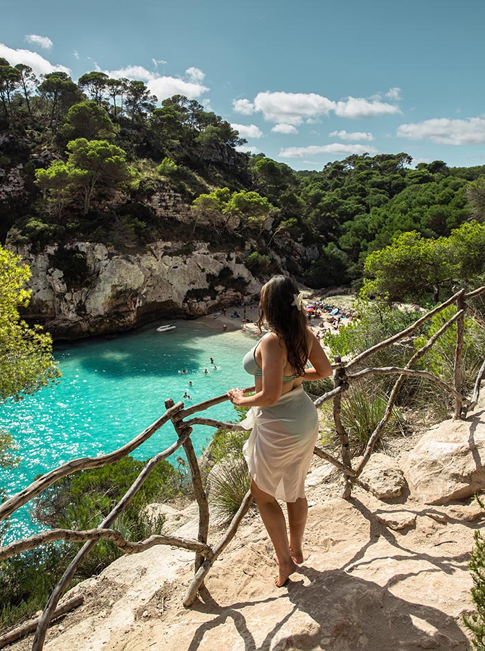 The image features a woman standing on a rocky path overlooking Macarelleta Beach in Menorca. She gazes out at the turquoise waters below, where swimmers enjoy the serene cove. The path is bordered by rustic wooden railings, and the surrounding landscape is lush with pine trees and greenery. The bright, sunny day highlights the vibrant colors of the beach, creating a picturesque view of this Mediterranean paradise.