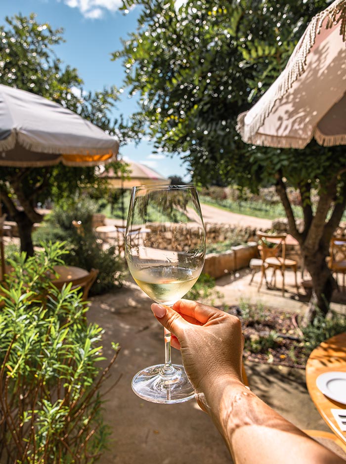 The image shows a hand holding a glass of white wine, raised against the backdrop of a sunny outdoor seating area at Bodegas Binifadet Winery in Menorca. The scene is framed by lush greenery, sun umbrellas, and wooden tables, creating a relaxed atmosphere for a wine-tasting experience. The bright sunlight filters through the leaves, highlighting the golden hue of the wine and the inviting vineyard setting beyond.