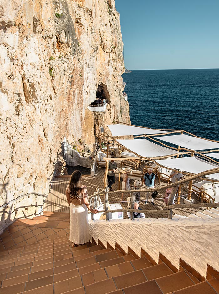 The image depicts a woman in a light-colored dress walking down terracotta steps towards an outdoor seating area at Cova d'en Xoroi, a cliffside bar in Menorca. The bar's seating is covered with white sunshades, with a wooden railing and beams adding to the rustic charm. The background reveals the expansive view of the Mediterranean Sea and the surrounding rocky cliff.