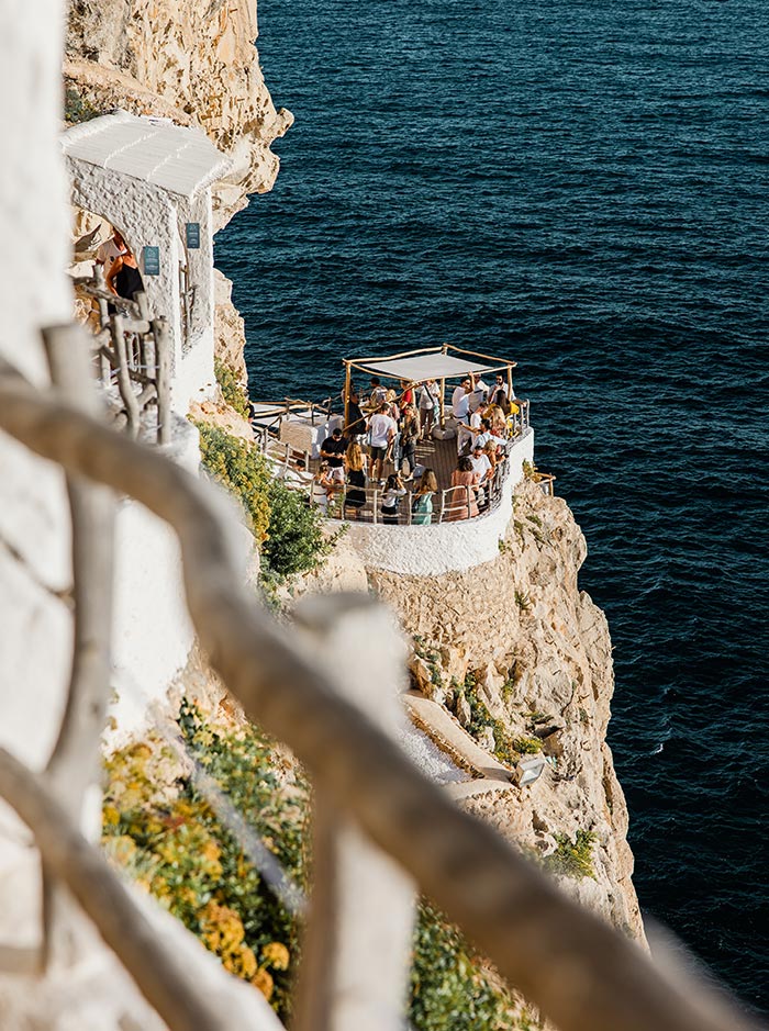 The image shows a lively gathering of people at an outdoor terrace of Cova d'en Xoroi, a cliffside bar overlooking the deep blue Mediterranean Sea. The terrace is built into the rocky cliff, featuring a wooden frame with a light canopy for shade. The vibrant scene captures visitors enjoying the unique views and atmosphere, with the steep cliffs and ocean waves below creating a dramatic backdrop.