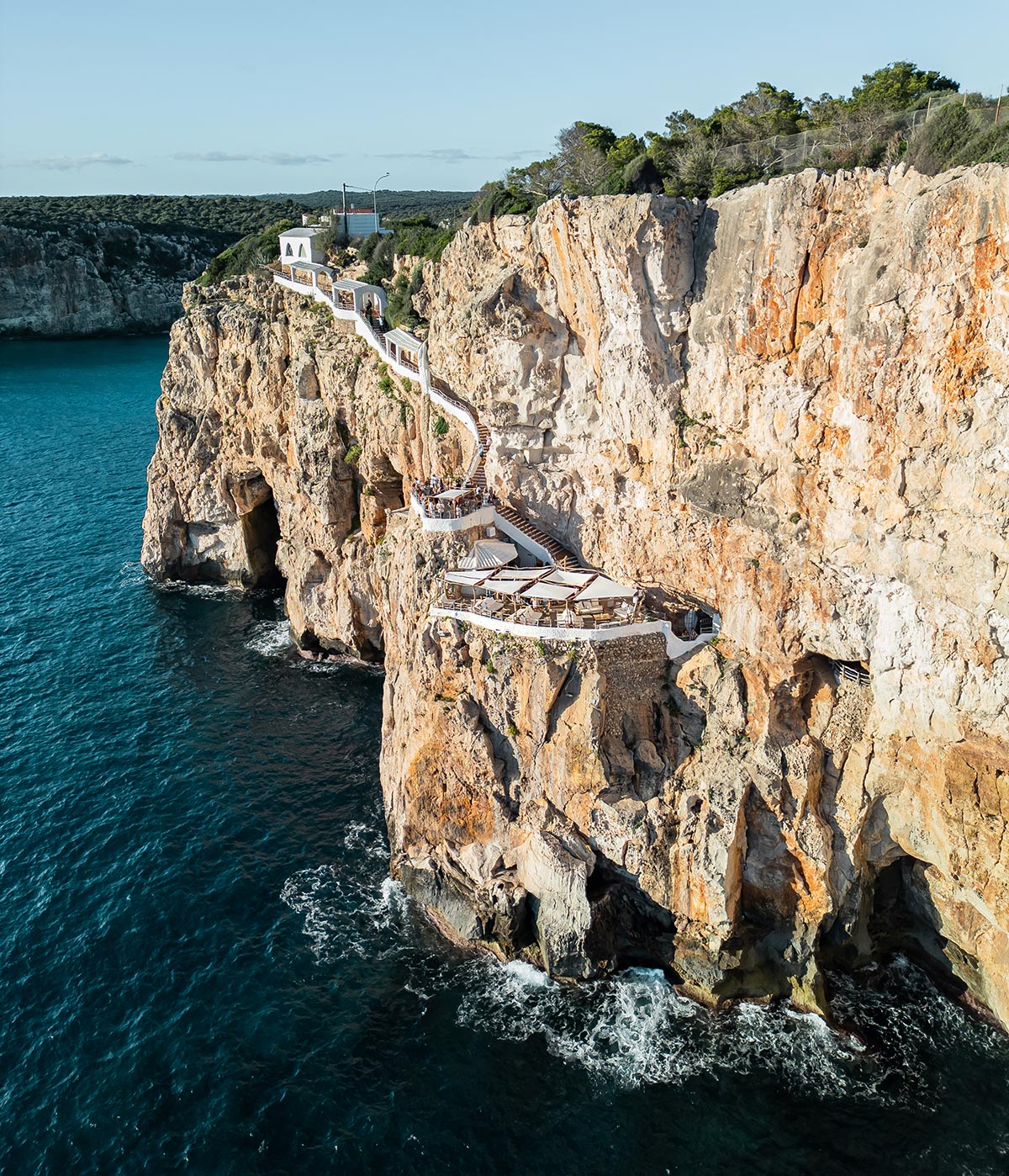 The image shows a scenic view of Cova d'en Xoroi, a cliffside bar perched on rugged cliffs above the turquoise Mediterranean Sea. The winding white staircases and seating areas are built into the natural rock formations, offering breathtaking panoramic views of the ocean. The sunlight illuminates the cliffs, enhancing their rich, earthy tones.