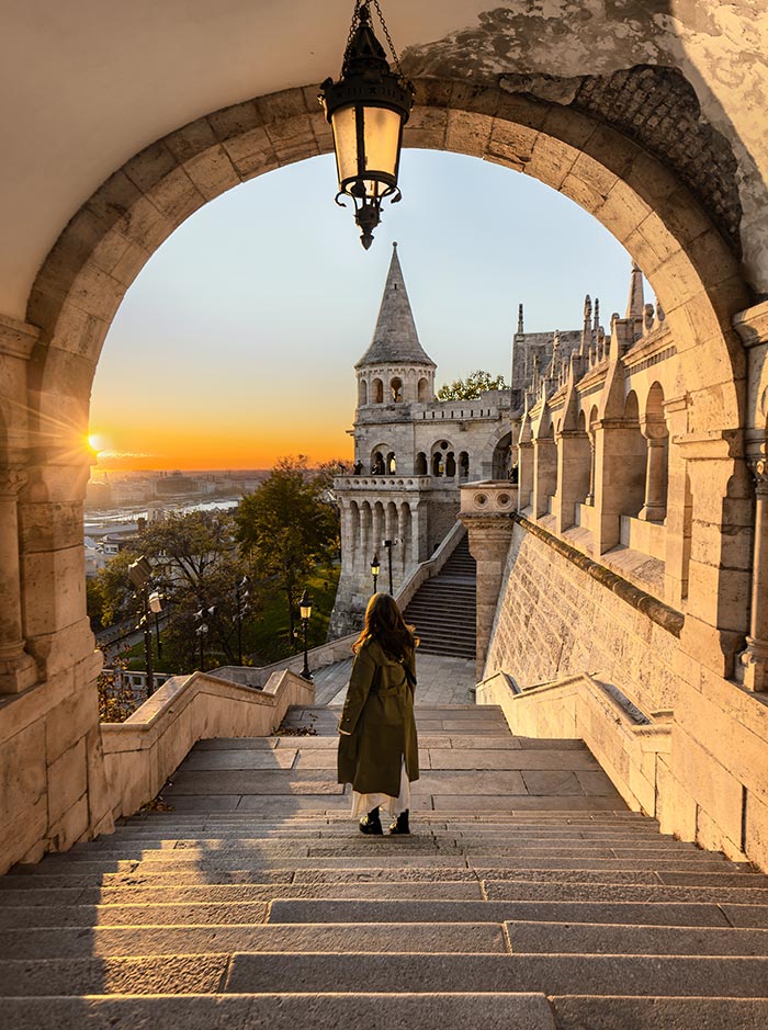 Alt text: A woman stands on the stone steps of Fisherman's Bastion in Budapest, Hungary, looking toward the sunrise. The sun casts a golden glow over the city and illuminates the iconic medieval-style towers and arches of the Bastion. A hanging lantern frames the scene, adding to the historic ambiance of the location.