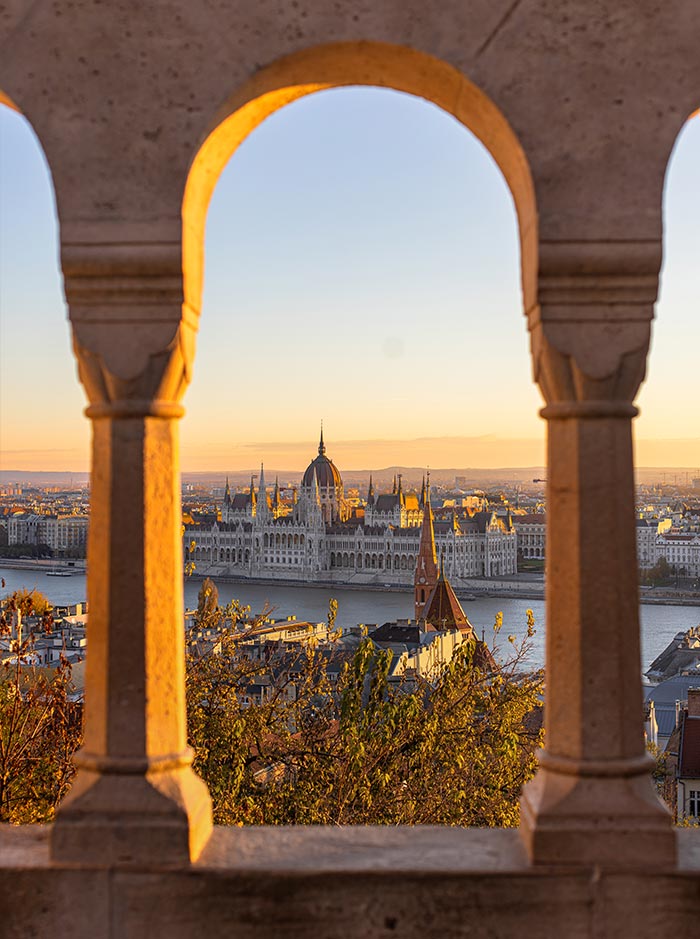 Alt text: The Hungarian Parliament Building in Budapest is framed by the arched stone windows of Fisherman's Bastion. The warm, golden light of sunrise highlights the intricate Gothic Revival architecture of the Parliament, with the Danube River flowing beside it. This scenic view captures the contrast between historic stonework and the grandeur of the cityscape.