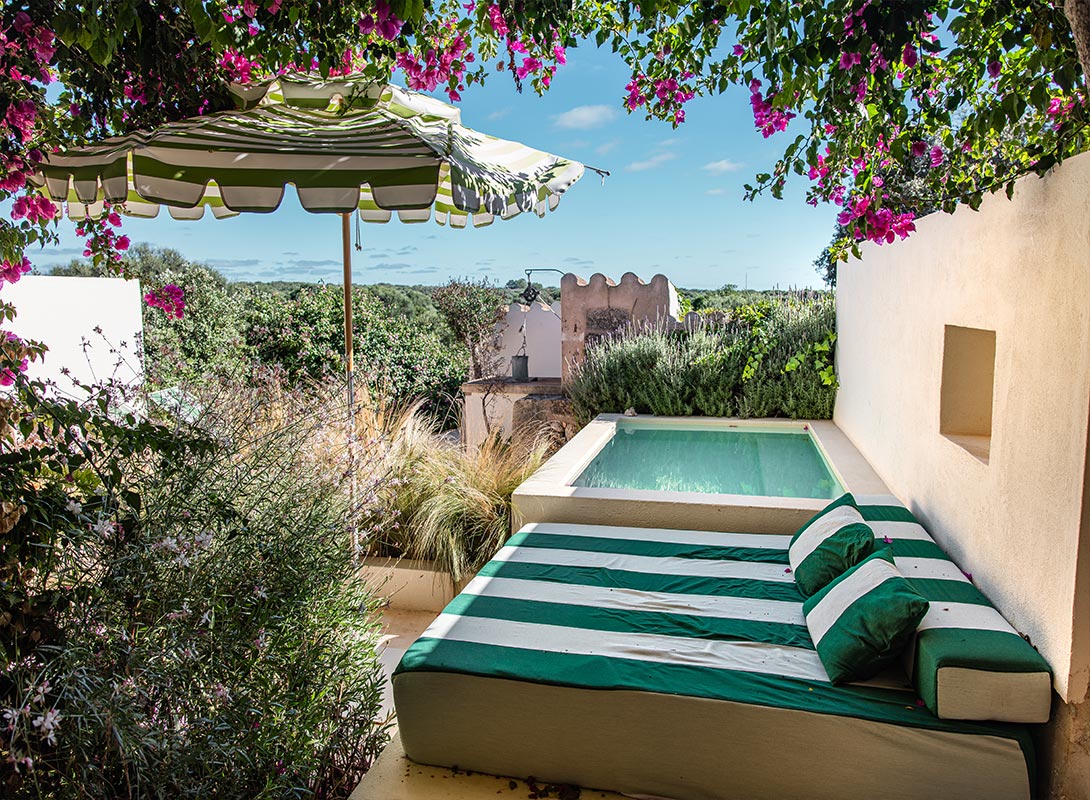 The image showcases a serene outdoor nook at Morvedra Nou in Menorca, featuring a cozy lounging area with a green and white striped daybed beside a small private plunge pool. Above, a matching green and white striped umbrella provides shade, while vibrant pink bougainvillea flowers drape overhead, adding a pop of color. The surrounding lush garden and open countryside beyond create a peaceful and secluded atmosphere, perfect for relaxation under the Mediterranean sun.