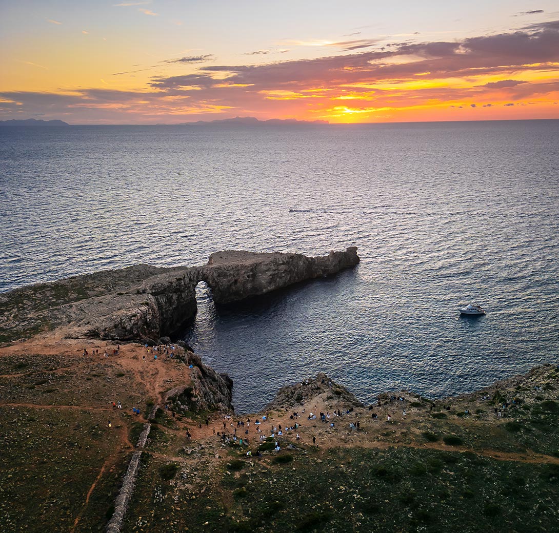 The image captures a stunning sunset view at Pont d'en Gil in Menorca, where the sun sets over the Mediterranean Sea, painting the sky in shades of orange, pink, and purple. The natural rock arch extends into the water, creating a dramatic coastal scene. Below, small groups of people can be seen gathered along the rocky cliffs, enjoying the serene atmosphere. A boat floats nearby, completing this picturesque seaside landscape.