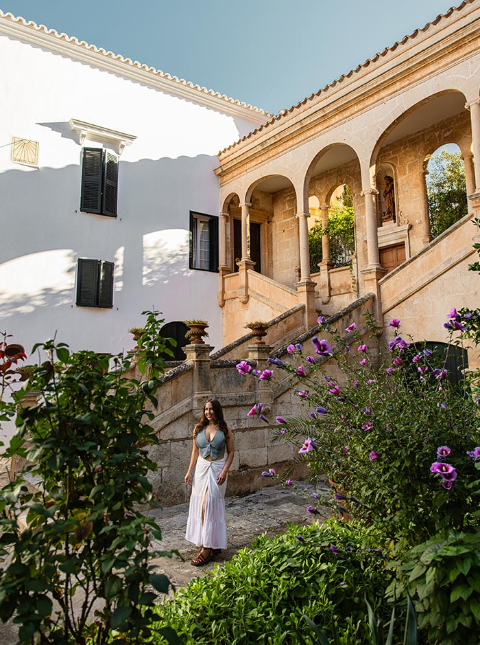 The image showcases the courtyard of the Bishop's Palace in Ciutadella, Menorca. A woman in a flowing white skirt and a light top strolls through a garden filled with blooming purple flowers. The scene is framed by an elegant stone staircase leading to an arched terrace, featuring classic Mediterranean architecture with warm sandstone tones. The sunlight highlights the details of the building's arches and lush greenery, creating a peaceful and picturesque atmosphere.