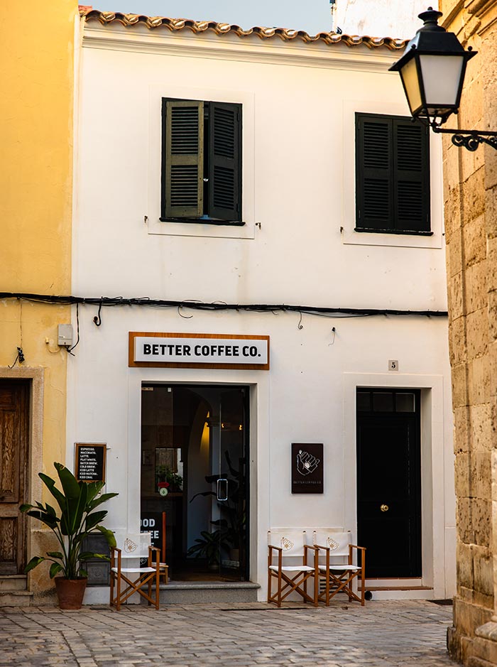 The image shows the exterior of Better Coffee Co., a charming coffee shop in Ciutadella, Menorca. The café is housed in a whitewashed building with dark green shutters, set against a cobblestone street. The entrance features a wooden-framed sign, potted plants, and a pair of director's chairs for outdoor seating. A classic black lantern adds to the quaint, inviting atmosphere of this cozy corner spot.