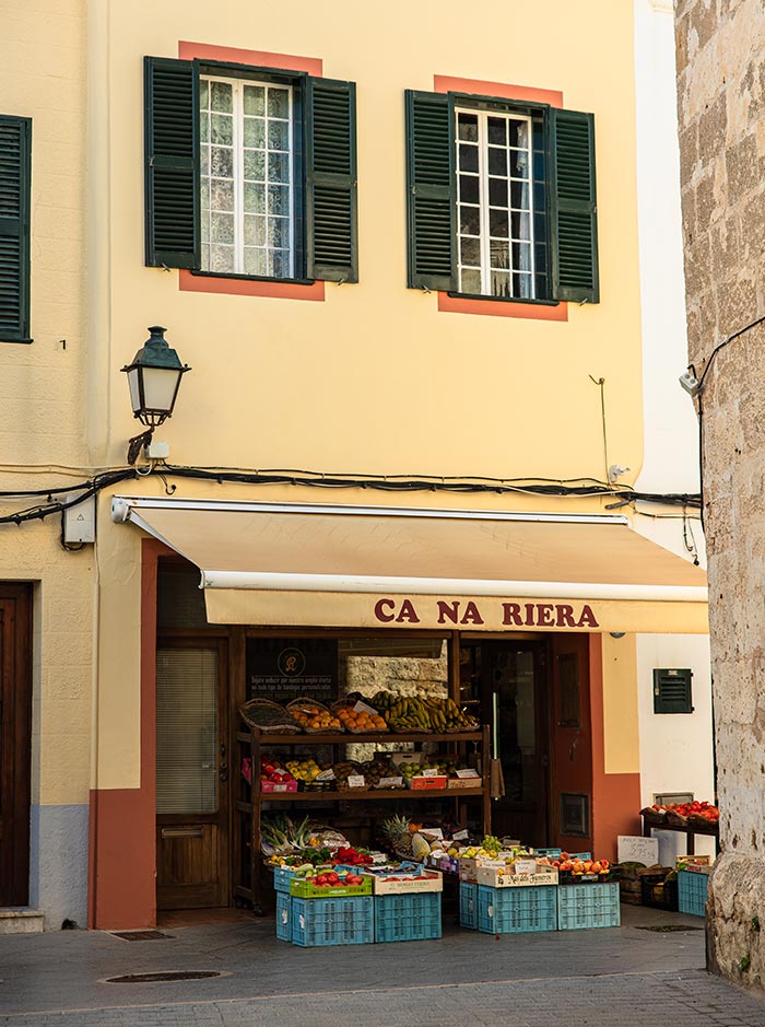 The image shows the exterior of Ca Na Riera, a quaint market shop in Ciutadella, Menorca. The building is painted a soft yellow with red accents around the windows, which have dark green shutters. Under a beige awning, the shop displays a colorful assortment of fresh fruits and vegetables in wooden crates and baskets, inviting passersby to browse the local produce. A classic lantern hangs beside the entrance, adding to the traditional charm of this corner market.