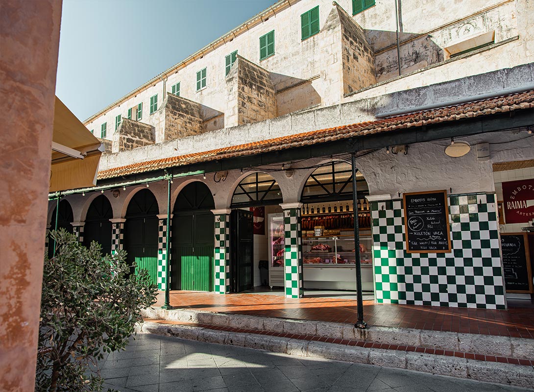 The image captures a traditional market building in Ciutadella, Menorca. The exterior features green and white checkered tiles, arched doorways, and large green shutters, creating a classic Mediterranean aesthetic. Inside, a market stall displays cured meats and other local delicacies. The scene is bathed in warm sunlight, with shadows casting patterns on the red-tiled floor, highlighting the vibrant atmosphere of this historic market area.