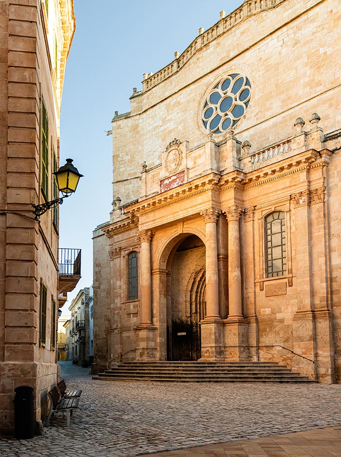 The image captures a peaceful street scene in Ciutadella, Menorca, featuring a grand stone church with a rose window and elegant columns at its entrance. The warm, golden light of the sun illuminates the intricate stonework, highlighting the historic architecture. A cobblestone street leads to the church, with a classic lantern and a wooden bench adding to the old-world charm of the surroundings. The scene is serene, reflecting the timeless beauty of the town.