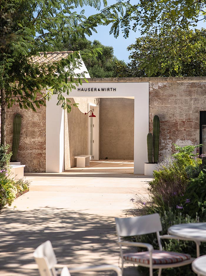 The image shows the entrance to the Hauser & Wirth gallery in Menorca, framed by a modern white archway set against a rustic stone wall. The gallery's name is displayed above the entrance, and potted cacti flank the pathway, adding a touch of greenery. The surrounding area includes lush plants and trees, creating a serene atmosphere that blends art and nature. A small seating area with a white table and chairs is visible in the foreground.