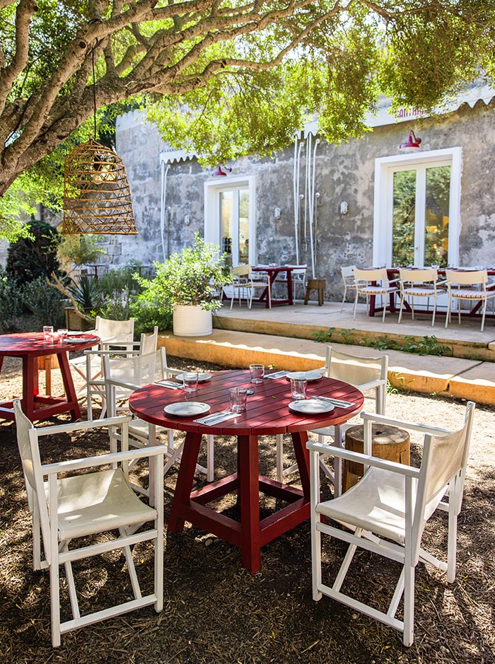 The image features an outdoor dining area at Hauser & Wirth Menorca, with red wooden tables and white chairs arranged under the shade of a large, leafy tree. The setup includes neatly set plates and glasses, ready for guests. The rustic building in the background has a textured stone façade, with large windows and a few colorful decorations, creating a relaxed, charming ambiance amidst the lush greenery.