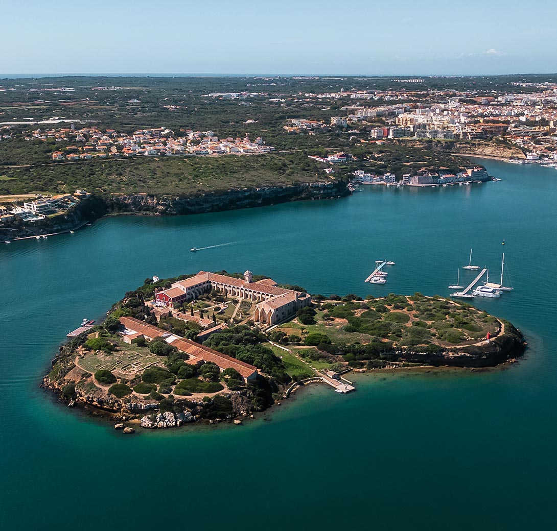 The image displays an aerial view of Hauser & Wirth Menorca, an art center situated on an island surrounded by the calm, turquoise waters of Mahón's harbor. The complex includes historic buildings, beautifully restored, with gardens and walkways that blend with the natural landscape. In the background, the main town of Mahón can be seen along the coastline, with a few yachts anchored near the island, emphasizing the serene and picturesque setting.