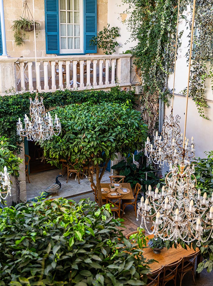 The image captures a cozy outdoor seating area at the Jardí de ses Bruixes Café in Mahón, Menorca. The space is filled with lush greenery, including trees that provide shade to wooden tables and chairs. Elegant chandeliers hang from above, adding a touch of sophistication to the rustic courtyard setting. A balcony with blue shutters overlooks the garden, enhancing the charming and intimate ambiance of this hidden café.