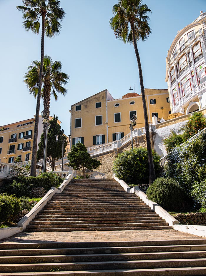 The image depicts a grand stone staircase leading up to colorful historic buildings in Mahón, Menorca. Tall palm trees line the steps, adding a tropical touch to the scene, while lush greenery surrounds the path. The warm yellow and earthy tones of the buildings, coupled with the bright blue sky, create a vibrant and inviting Mediterranean atmosphere.