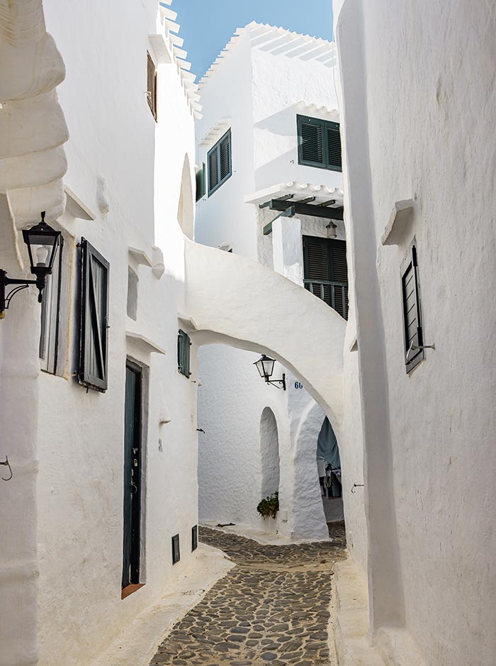 The image showcases a narrow, winding street in Binibeca Vell, a charming fishing village in Menorca. The path is lined with traditional whitewashed buildings, featuring black wooden shutters and small windows. An arched walkway connects the buildings, adding to the quaint and cozy atmosphere of the alley. The soft sunlight brightens the scene, emphasizing the clean, minimalist Mediterranean architecture.