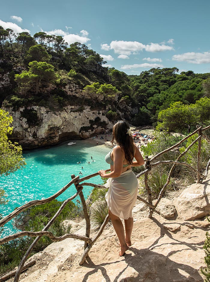 The image features a woman standing on a rocky path overlooking Macarelleta Beach in Menorca. She gazes out at the turquoise waters below, where swimmers enjoy the serene cove. The path is bordered by rustic wooden railings, and the surrounding landscape is lush with pine trees and greenery. The bright, sunny day highlights the vibrant colors of the beach, creating a picturesque view of this Mediterranean paradise.