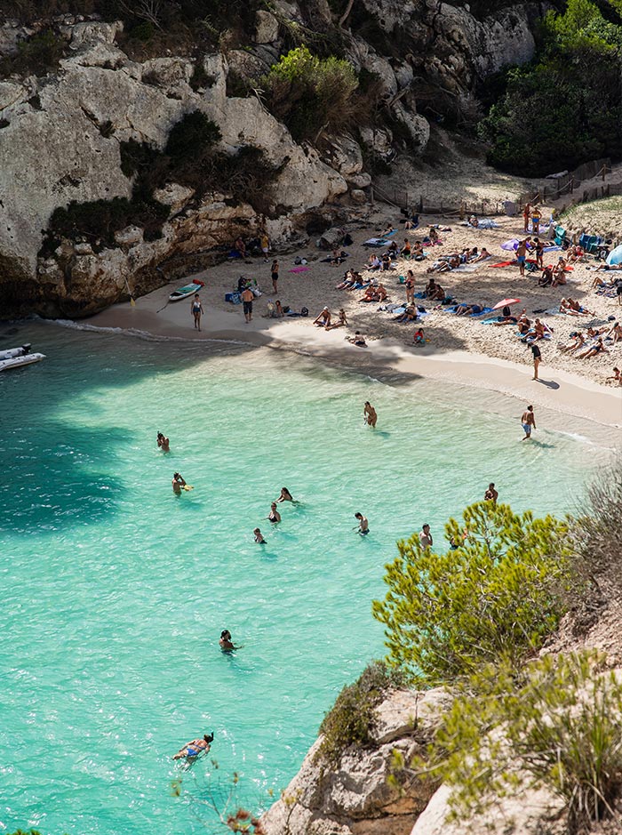 The image captures a closer view of Macarelleta Beach in Menorca, highlighting the clear turquoise water where people are swimming and enjoying the refreshing sea. The sandy shore is lined with sunbathers and colorful umbrellas, while the rocky cliffs and lush vegetation frame the cove. The sunlight dances on the water, creating a serene and inviting atmosphere, perfect for a day of relaxation in this beautiful Mediterranean setting.