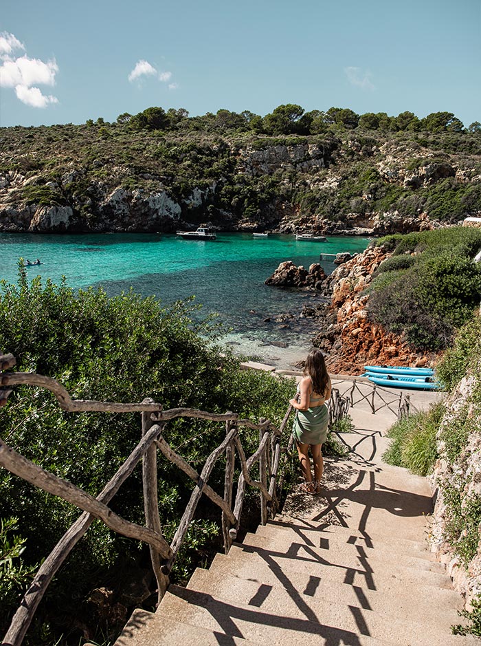 The image shows a woman descending a set of stone steps towards the turquoise waters of Cala Es Canutells in Menorca. The steps are lined with rustic wooden railings, surrounded by lush greenery and rocky terrain. In the background, small boats float in the clear, calm cove, with the rugged cliffs and verdant hills framing the serene beach scene. A bright, sunny day enhances the natural beauty of this peaceful coastal spot.