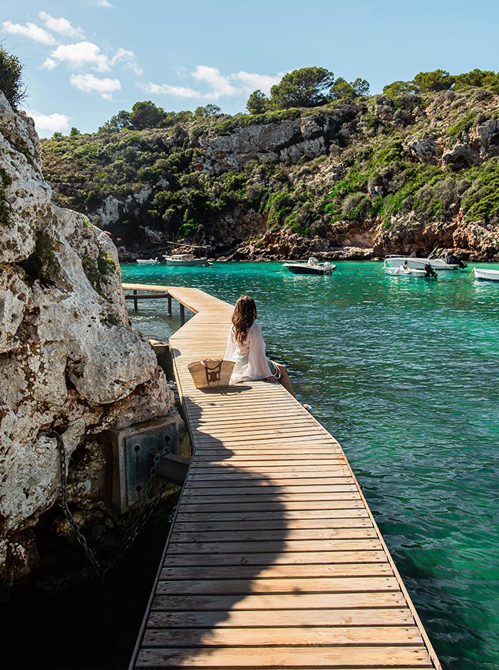 The image features a serene scene at Cala Es Canutells in Menorca, with a woman sitting on a wooden boardwalk that extends over the turquoise waters of the cove. The boardwalk curves gently, leading towards the rocky cliffs covered in lush greenery. A few boats float in the calm water, adding to the tranquil atmosphere. The woman sits with a beach bag beside her, enjoying the peaceful surroundings and the bright, sunny day.