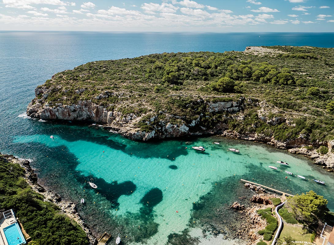 The image shows a beautiful aerial view of Cala Es Canutells, a small cove in Menorca. The turquoise waters of the bay are crystal clear, gradually deepening to a rich blue as they meet the open sea. A few boats are anchored in the calm water, while lush greenery covers the surrounding rocky cliffs. The cove's secluded and serene atmosphere is perfect for a relaxing day by the water, under the sunny Mediterranean sky.