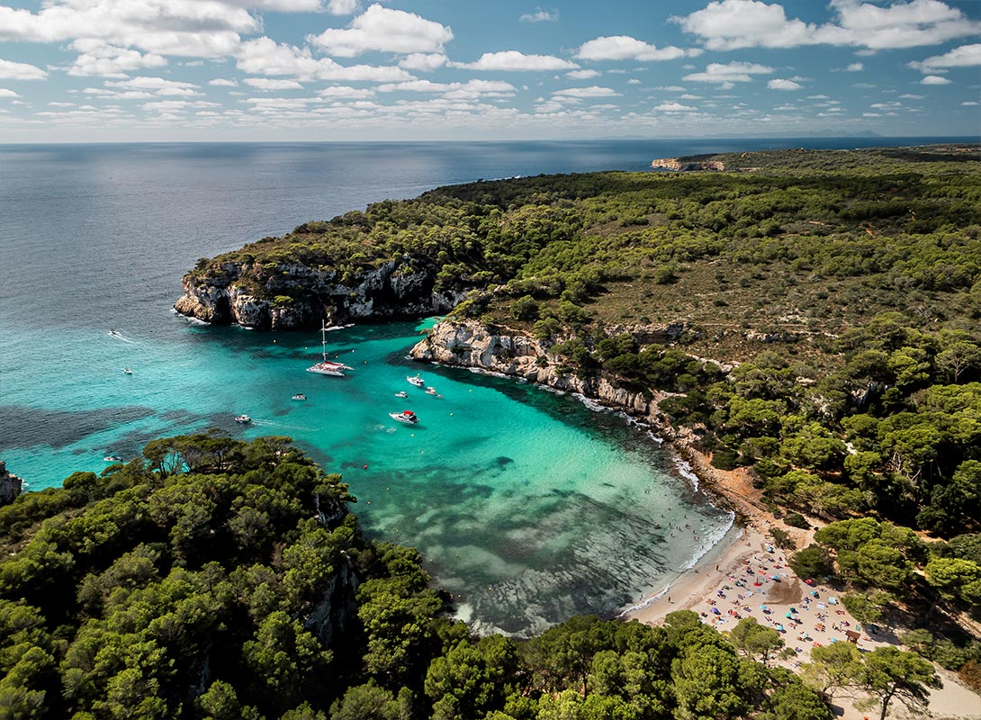 The image shows an aerial view of Cala Macarella, a stunning beach in Menorca. The cove is surrounded by rocky cliffs and dense green forest, with crystal-clear turquoise waters flowing into the Mediterranean Sea. The beach itself is dotted with colorful umbrellas and sunbathers enjoying the sunny day. Several boats are anchored in the calm bay, adding to the idyllic and serene atmosphere of this beautiful coastal landscape.